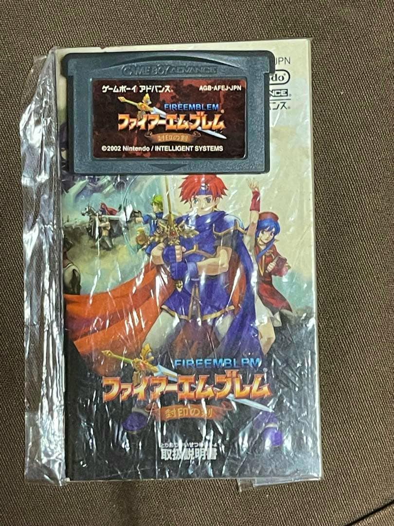 A Japanese Game Boy Advance cartridge for Fire Emblem shown on top of its cover art, both wrapped in plastic.