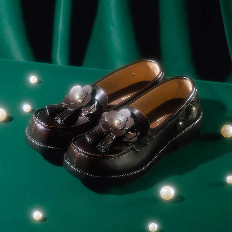 Pair of Fleur tassel leather loafers photographed on a green draped surface with scattered pearls.