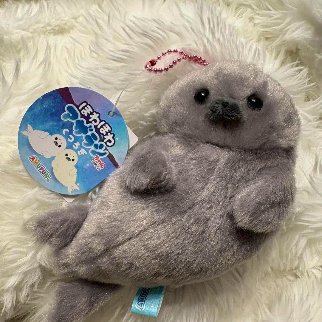 A gray plush seal toy lying on a white fluffy surface with a round product tag and pink ball chain attached.