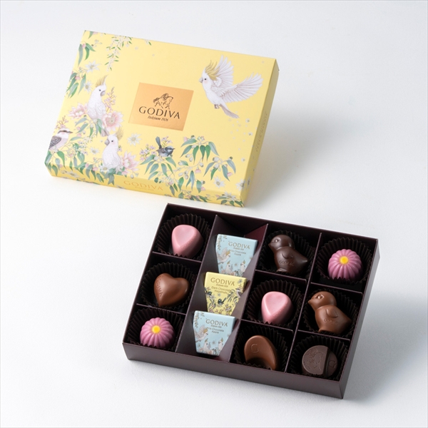 Open box of assorted chocolates in a compartment tray with the yellow decorative product box positioned behind it.