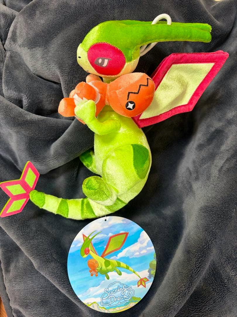 Green Flygon plush holding a small orange Nackler with a round "Soaring Dreams" tag, photographed on a dark blanket.