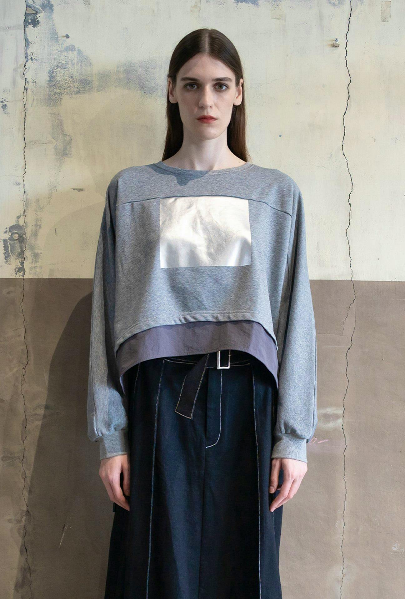 Model wearing a grey cotton dolman sweater with a rectangular silver foil print on the front.