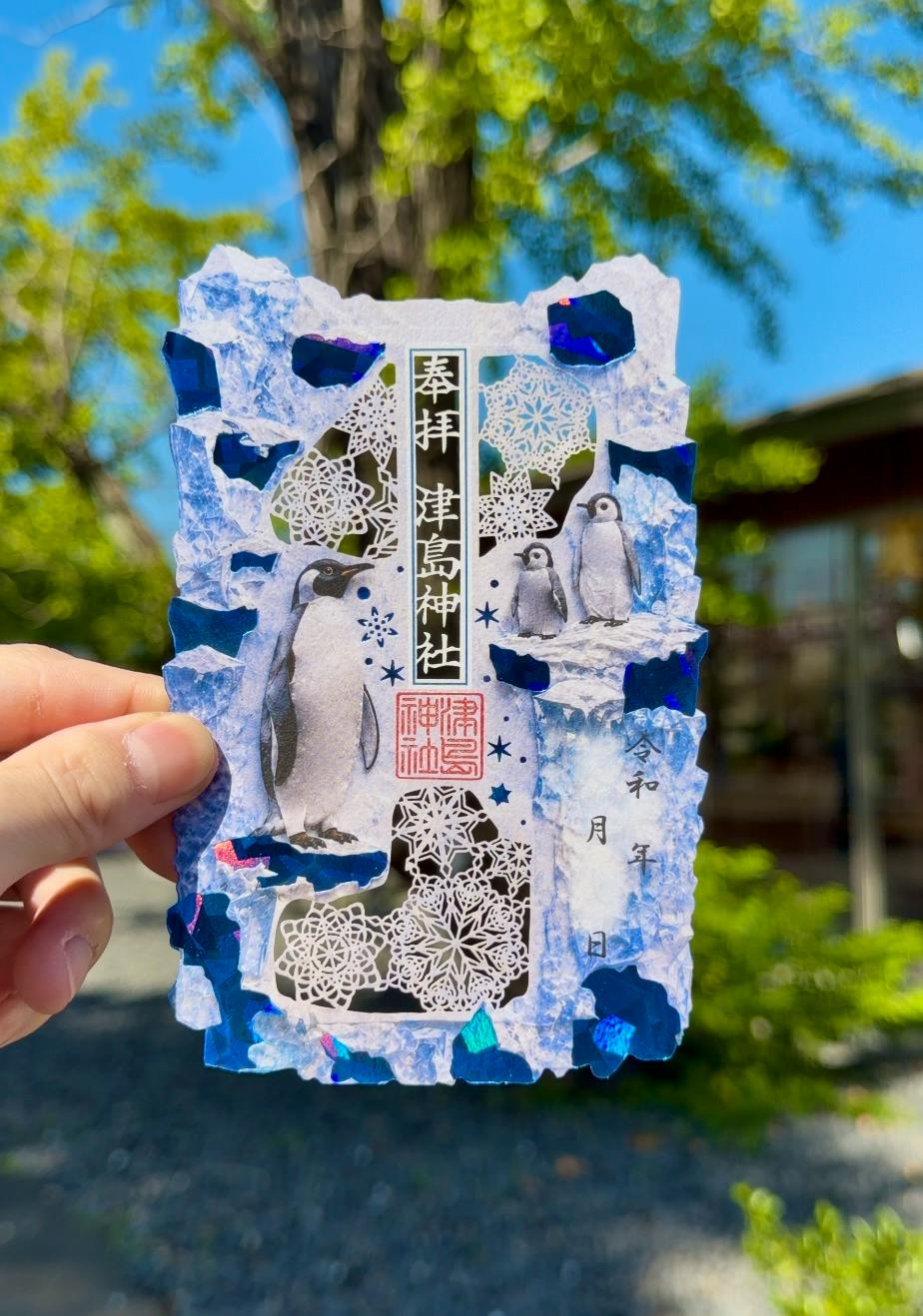 A hand holds a foil-stamped penguin-themed goshuin (shrine stamp) with blue torn edges, intricate paper cutouts, and red shrine seals against an outdoor background.