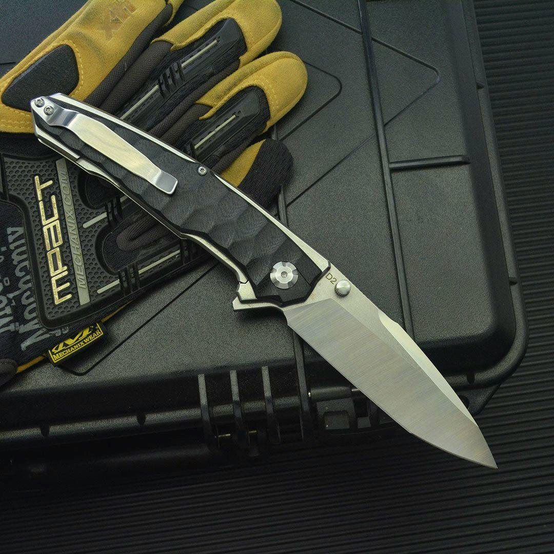 Open folding pocket knife with metal blade and textured handle resting on a black hard case beside gloves.
