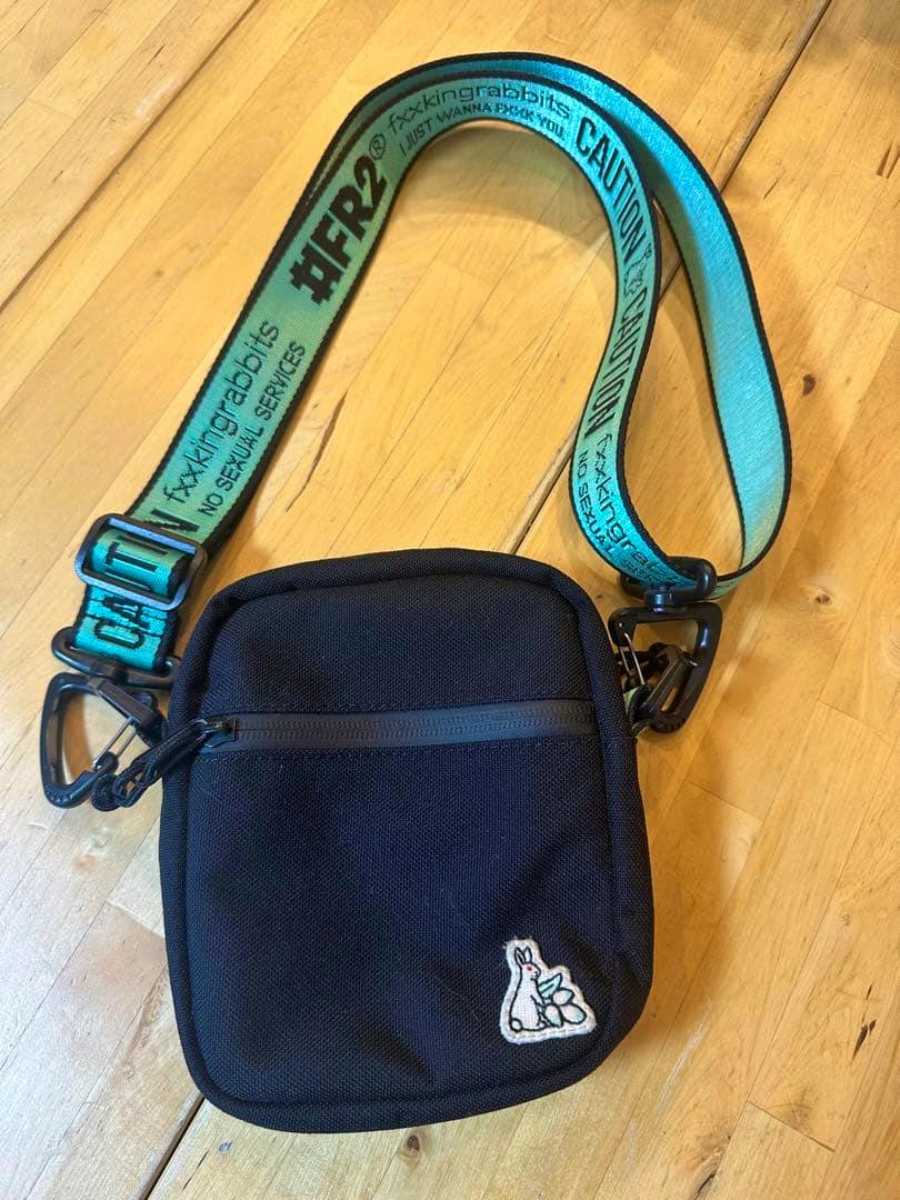 Black FR2 shoulder bag with a turquoise branded strap and a small embroidered rabbit patch, photographed on a wooden floor.
