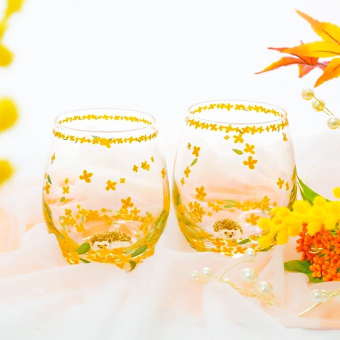 Two decorated drinking glasses with orange floral patterns and a small hedgehog illustration, displayed on a light pink fabric.