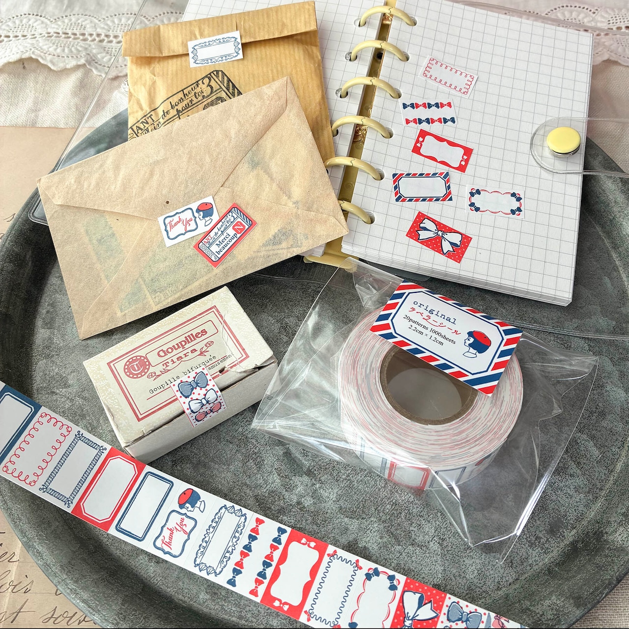 Photo showing the ラベラーシール「フレームリボン」 roll and strips of label stickers with sample stickers applied to envelopes and a notebook.