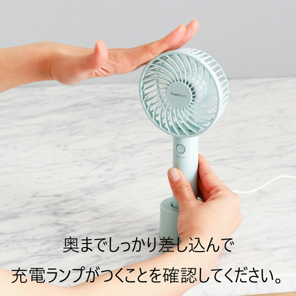 A white handheld fan being held by hands above its charging dock on a light surface with Japanese instructional text overlaid.