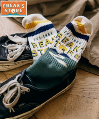 Men's jacquard tube socks with striped cuffs and multicolor jacquard logo pattern shown inside canvas sneakers on a wooden floor.