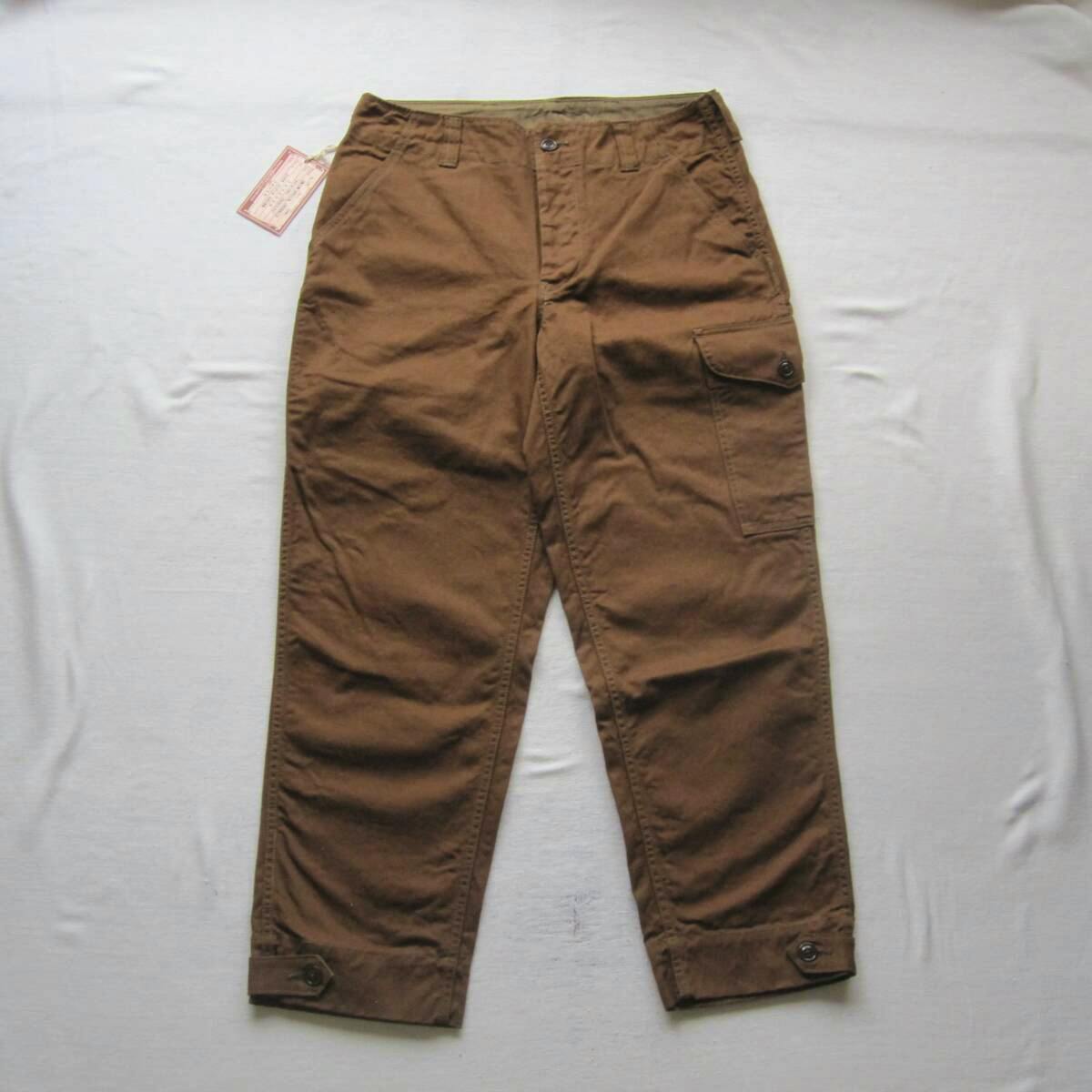 Brown mens cotton aviators' trousers laid flat showing the front with a cargo pocket and ankle tab details.