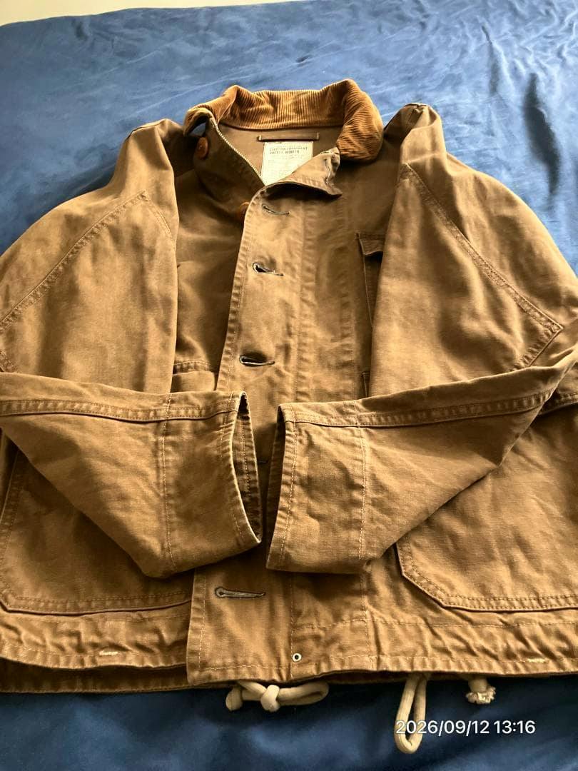 Flat-lay photo of a brown cotton men's military jacket (size 38) on a blue surface, showing the corduroy collar, front buttons, and drawstring hem.
