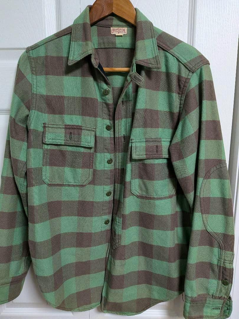 Front view of a men's cotton flannel checkered shirt in green and brown hanging on a wooden hanger.