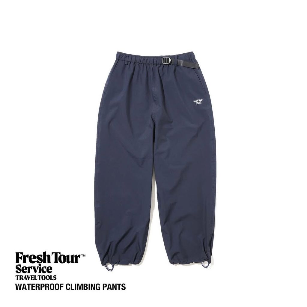 Flat-lay navy waterproof climbing pants (men's) on a white background, showing elastic cuffs, a buckle belt, and a small logo on the thigh.