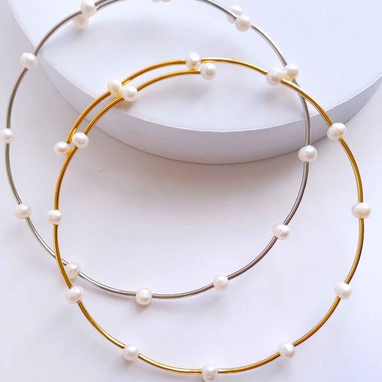 Two freshwater pearl line chokers, one gold-tone and one silver-tone, displayed on a white round platform.