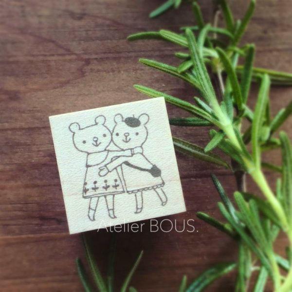 Wooden stamp showing an engraved illustration of two hugging bear children on a wooden block placed on a wooden surface with green sprigs alongside.