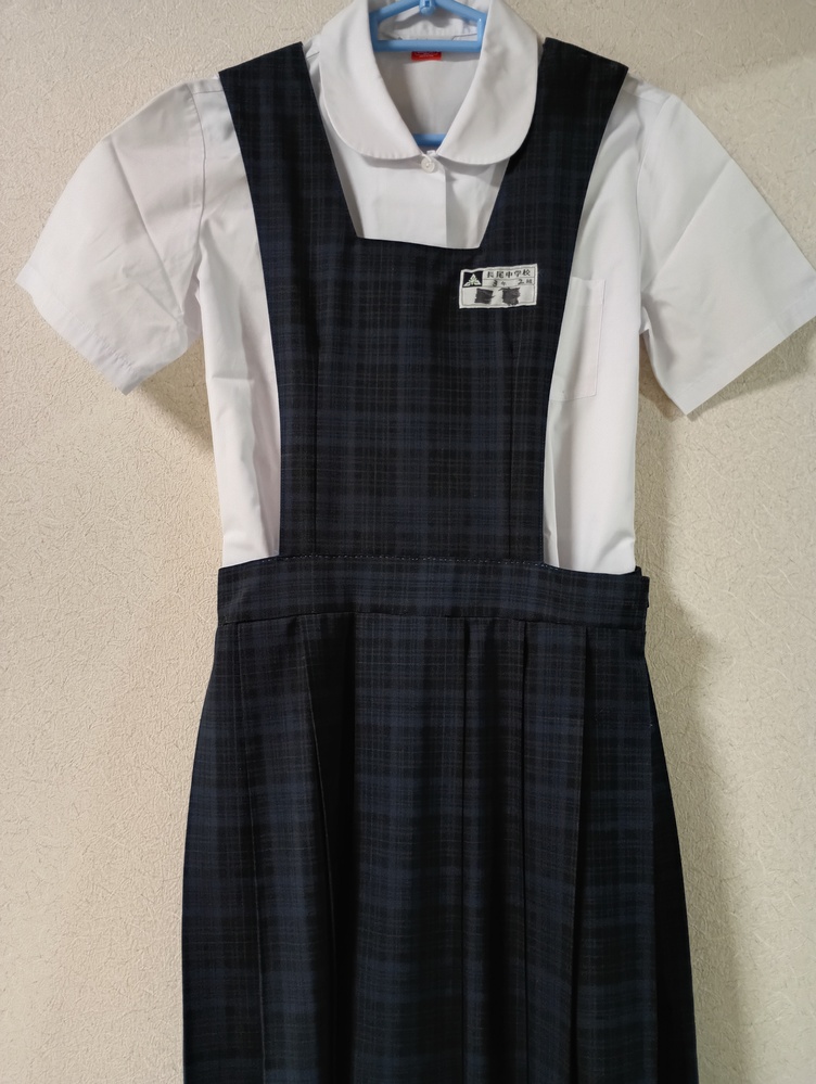 Front view of a women's summer school uniform set with a white short-sleeve blouse and a dark plaid pinafore hanging on a hanger.