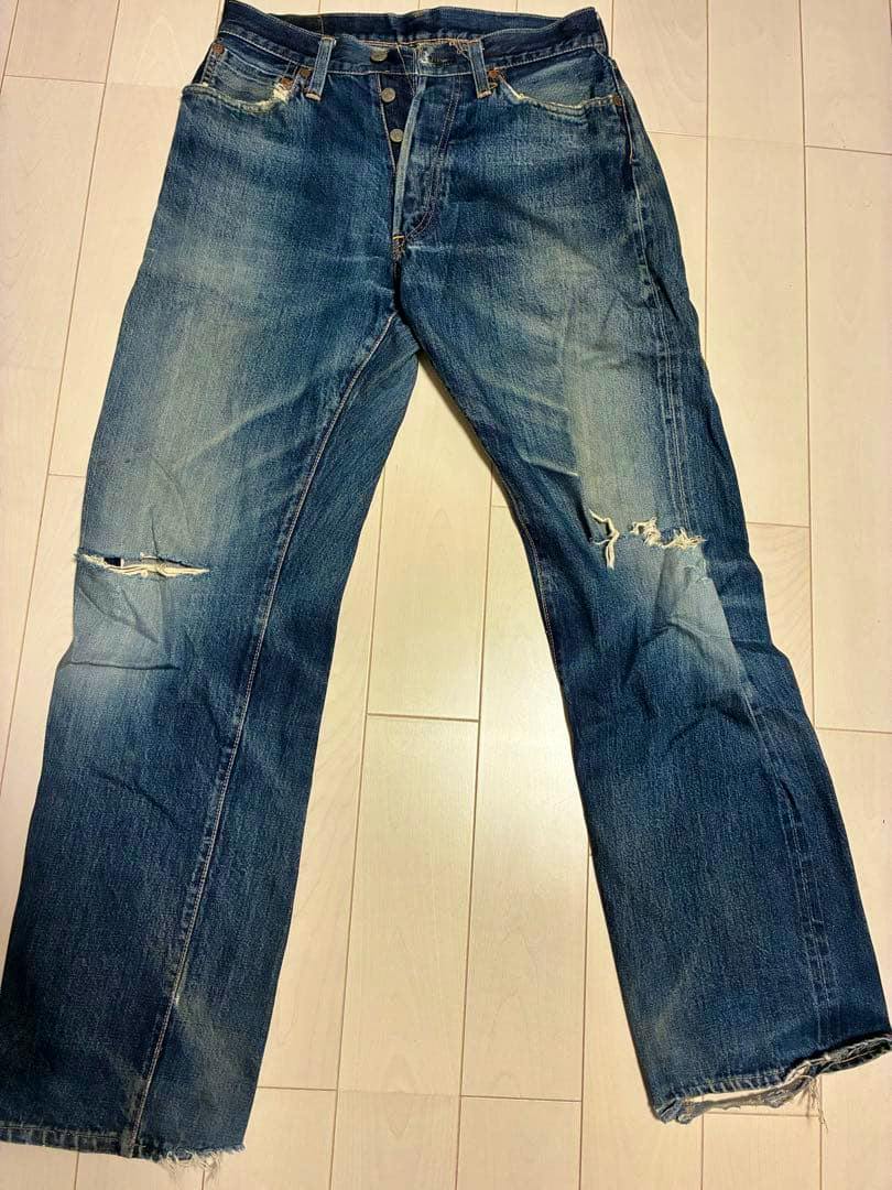 Front view of distressed denim jeans with visible button fly laid flat on a light wood floor.