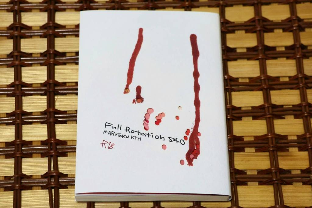 Photograph of the comic book "Full Rotation 540°" showing the white cover with red paint-like streaks, placed on a woven mat.