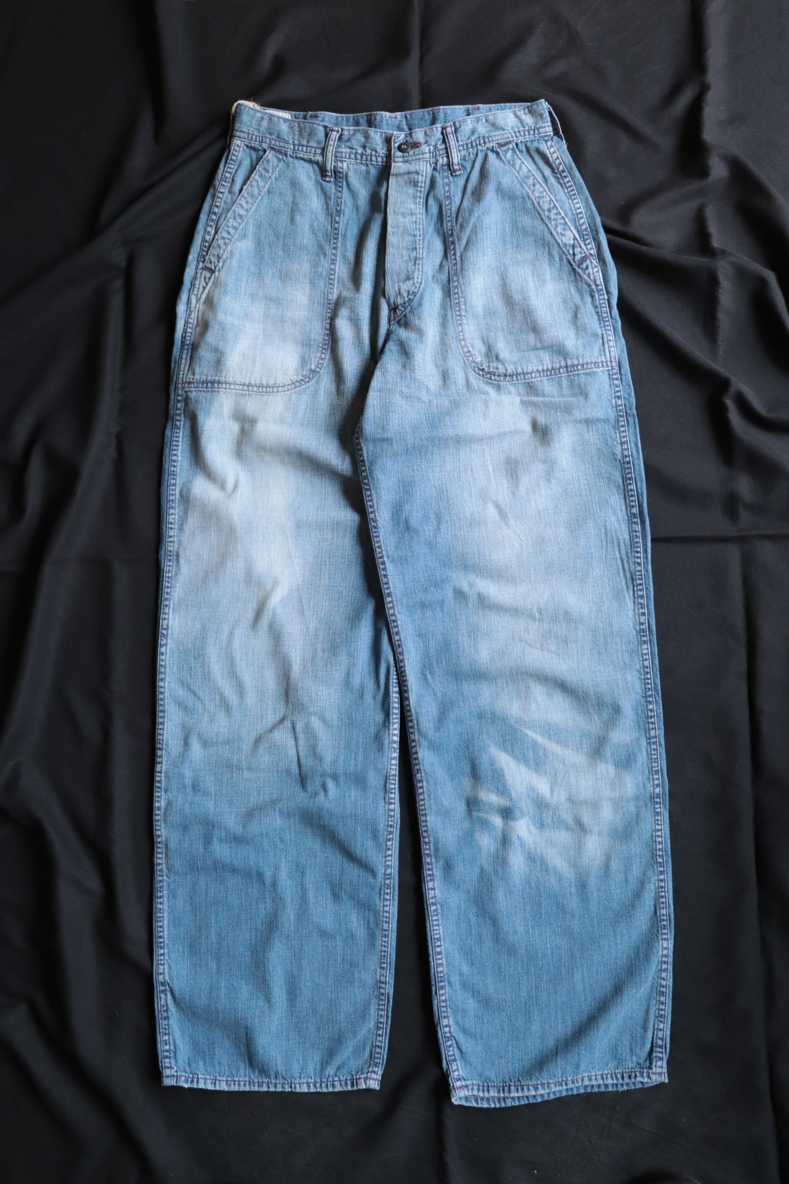 Front view of light-wash denim men's seamen's trousers laid flat on a dark background.