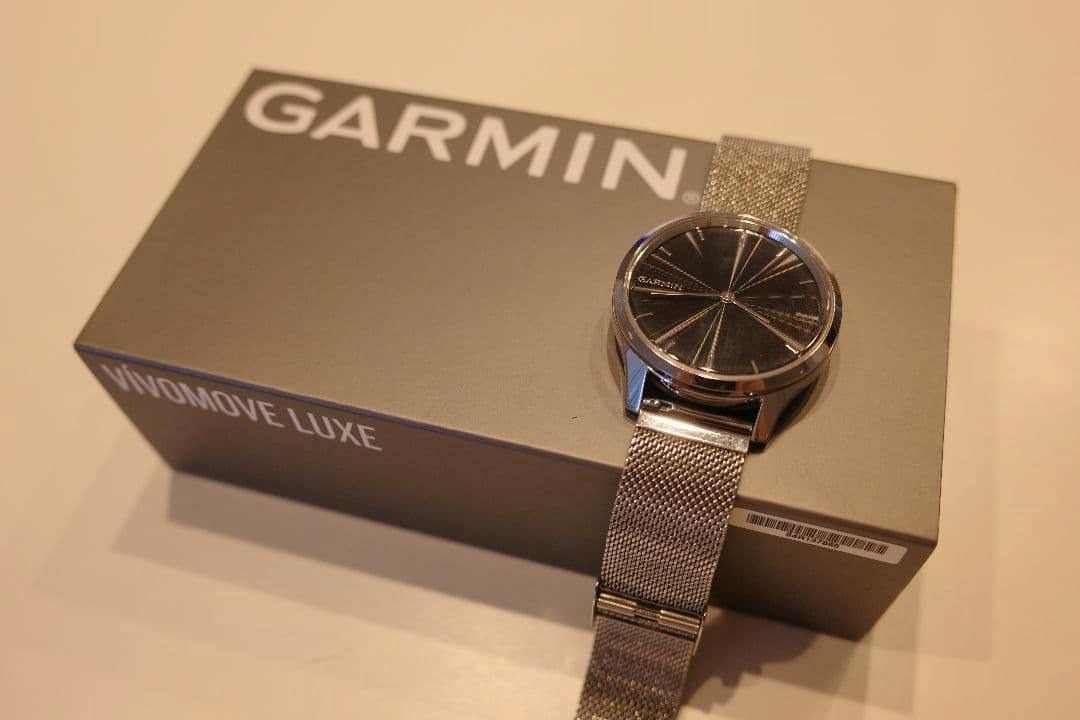 A Garmin VIVOMOVE LUXE watch with a Milanese mesh band resting on a Garmin-branded VIVOMOVE LUXE box.