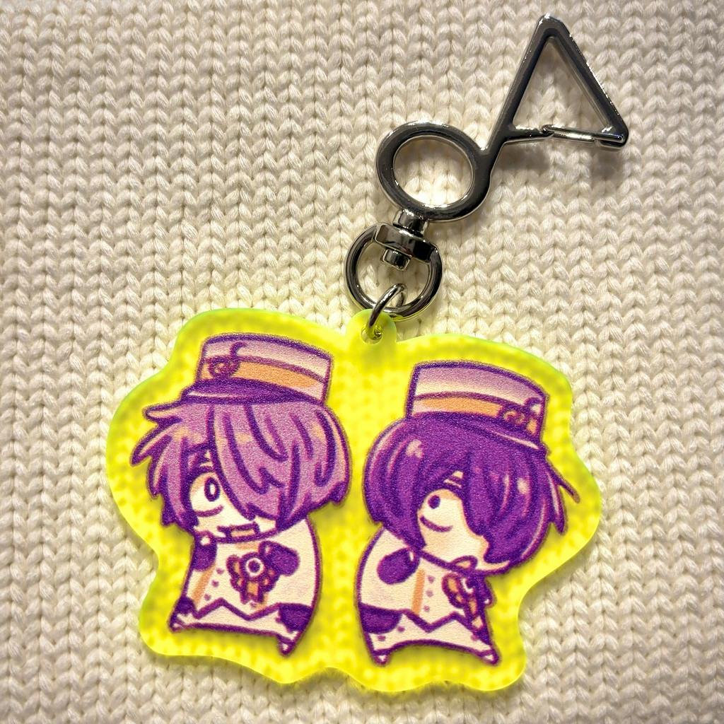 Photo of an acrylic keychain showing two chibi character illustrations on a yellow border with a metal clasp on a textured white surface.