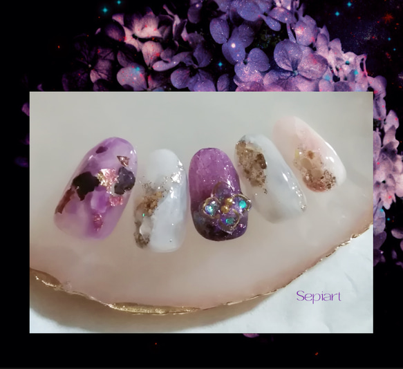 Five decorated gel nail tips with purple and white designs and 3D floral and rhinestone accents displayed on a shell.