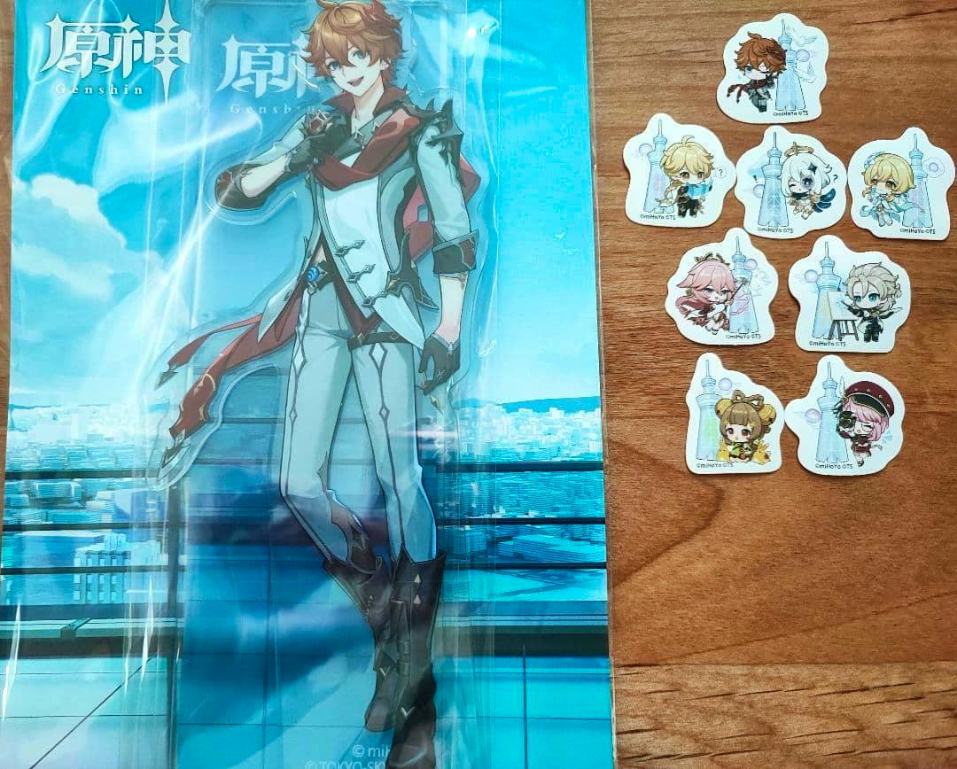 Packaged Tartaglia (タルタリヤ) acrylic stand from Genshin displayed next to several small chibi acrylic stickers on a wooden surface.