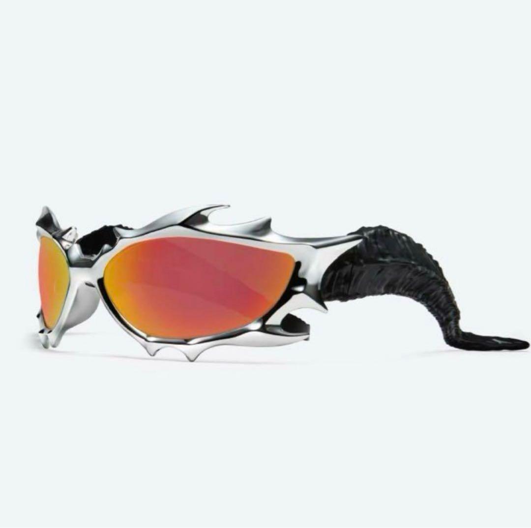 Gentle Monster sunglasses with a silver sculpted frame, orange mirrored lenses, and black textured tail-like temples on a white background.