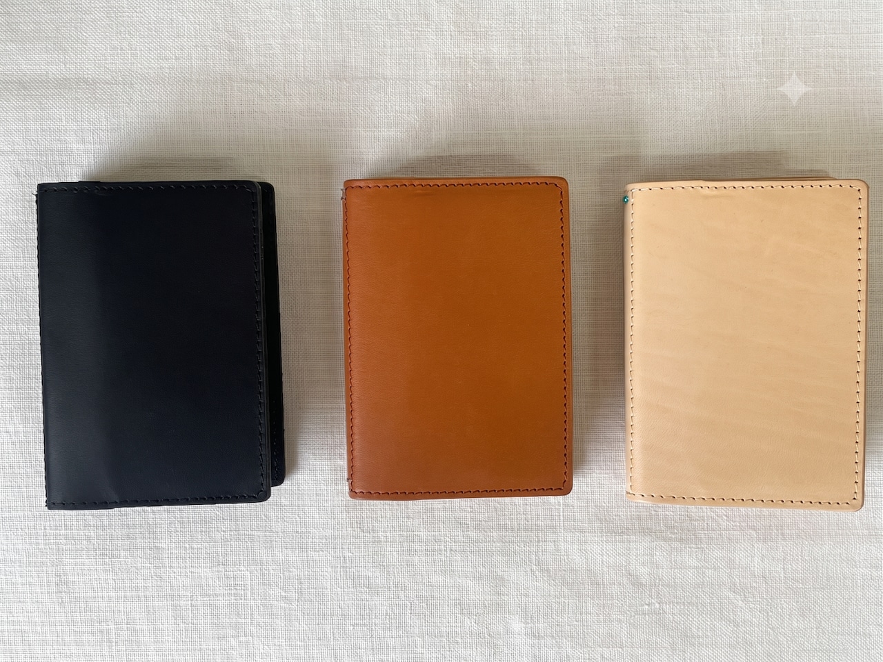 Three A7 leather notebook carry covers (black, brown, natural) laid side by side on a white fabric background.