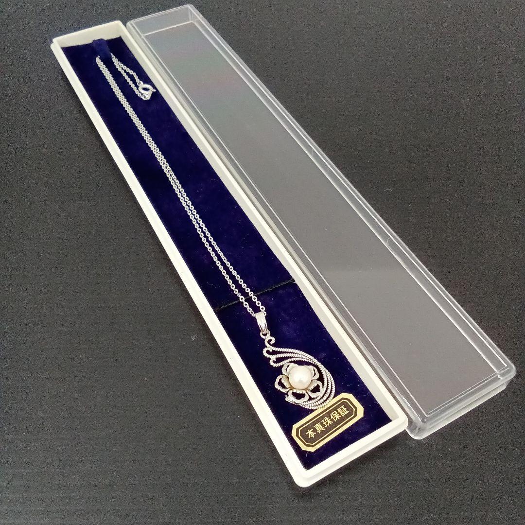 A pearl necklace with a silver pendant displayed inside a white rectangular presentation box with a navy velvet insert and a clear plastic lid.