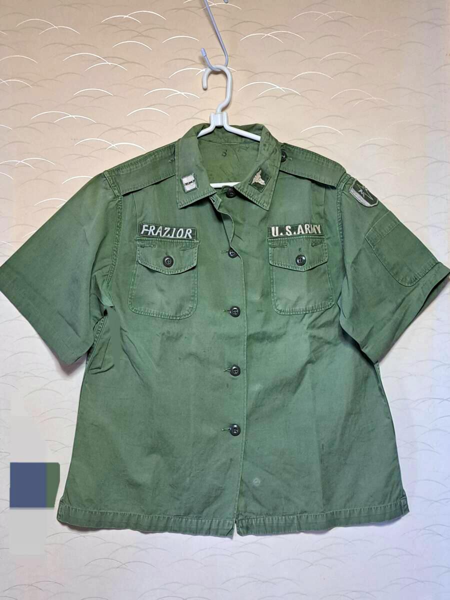 Front view of an olive-green men's cotton US Army OG-107 utility shirt with short sleeves, chest pockets, and name and service tapes, hanging on a hanger.
