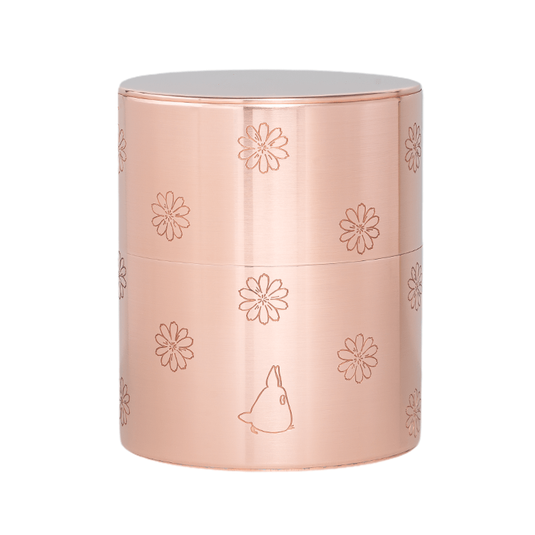 A metal cylindrical tea canister with engraved floral motifs and a small Totoro silhouette on the surface.