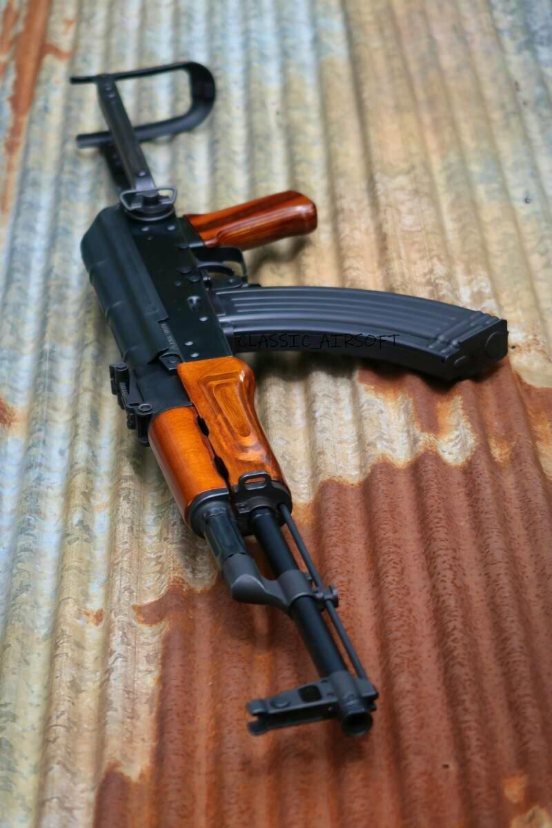 GHK AKMS airsoft-style rifle with wooden furniture and curved magazine on corrugated metal.