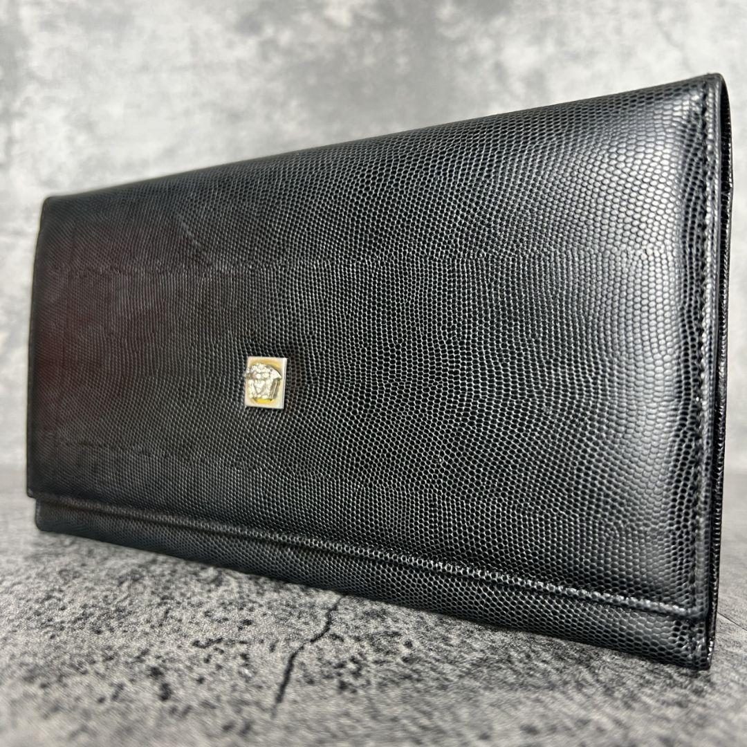 Black lizard-embossed long wallet (cow leather) with a small Medusa emblem, shown closed on a textured surface.