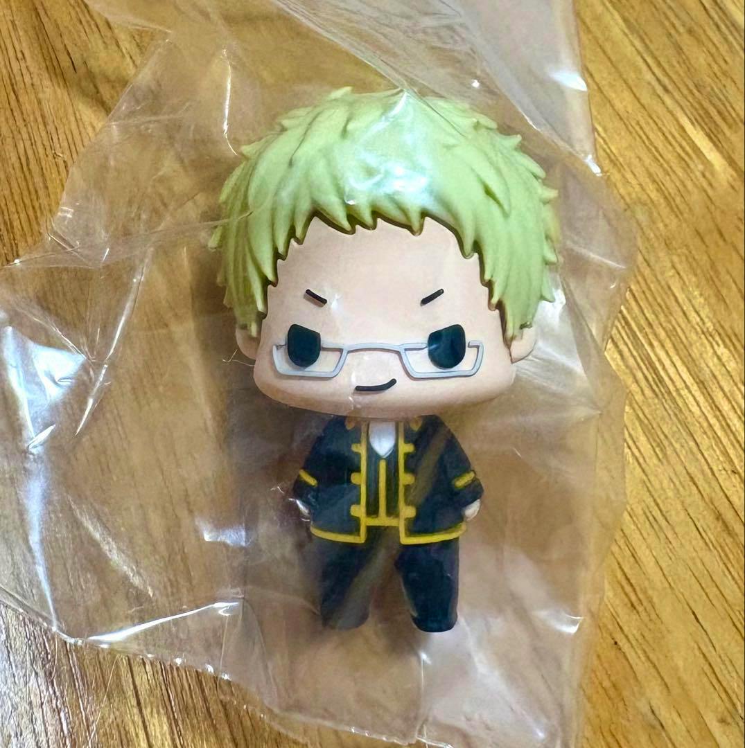 Chibi plastic figure from 銀魂 ちょこりんコレクション 伊東鴨太郎 in a clear plastic bag, showing green hair, glasses, and a dark outfit with yellow trim on a wooden surface.