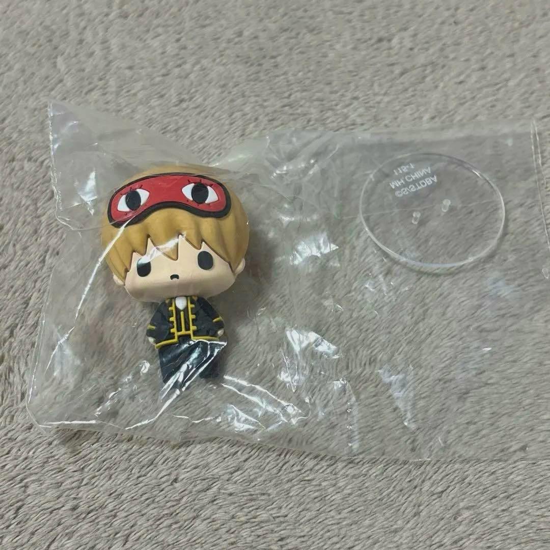 A small plastic 銀魂 ちょこりん mascot figure of 沖田総悟 in its clear bag with a round clear display stand on a beige surface.