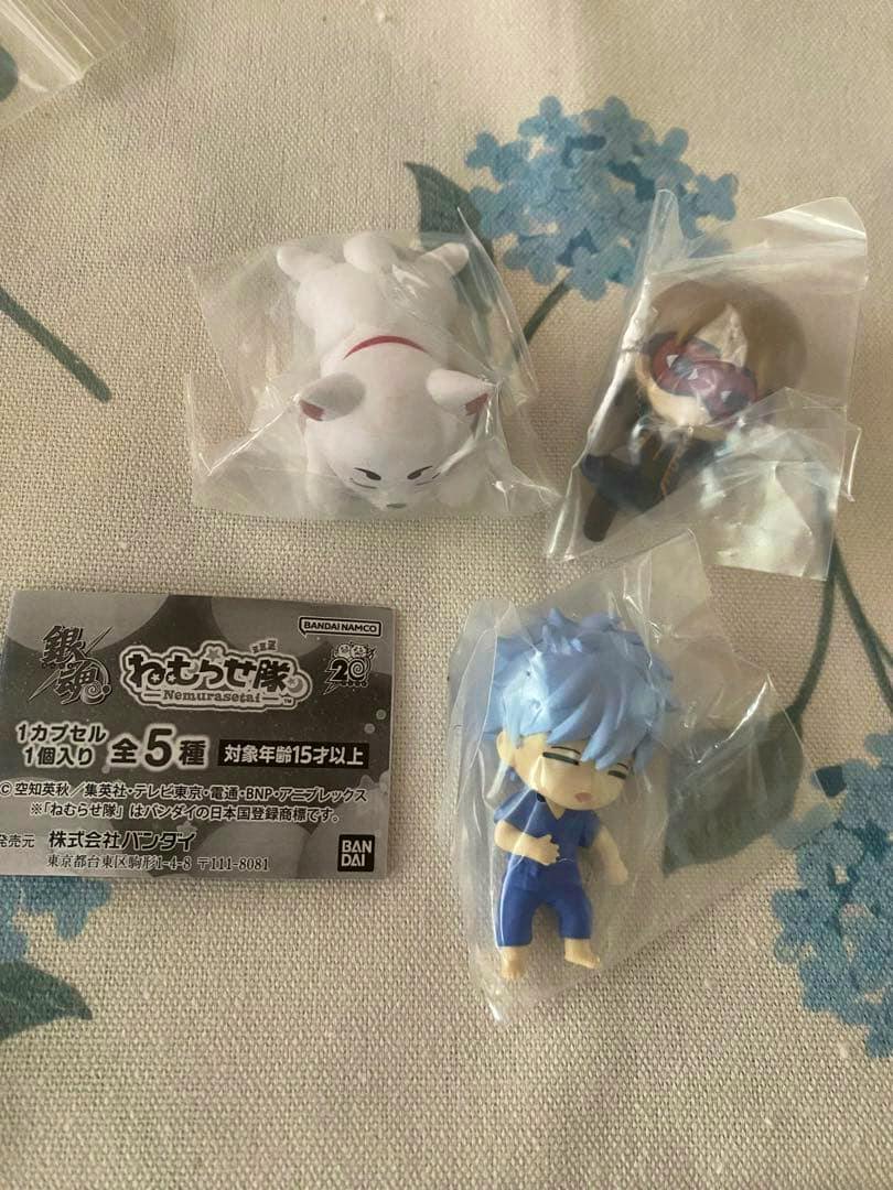 Three small Gintama Nemurasetai gacha plastic figures in individual clear bags with a Bandai product insert on a fabric background.