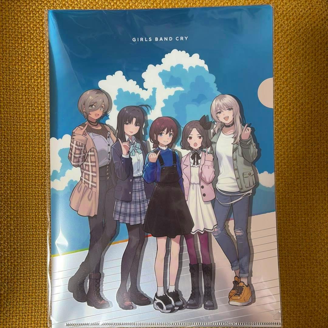 Photograph of a metal clear file showing five anime-style girls standing against a blue sky with clouds.