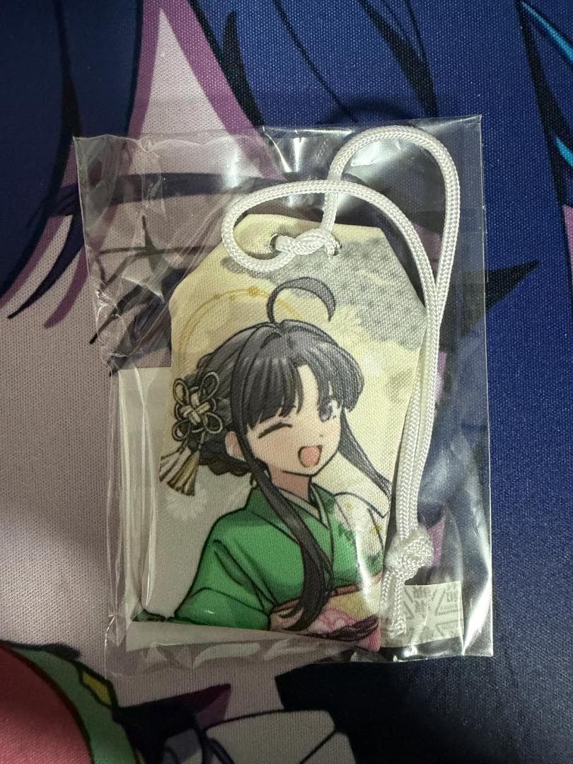 Unopened cloth amulet (omamori) in clear plastic packaging showing a smiling illustrated character and a white hanging cord.