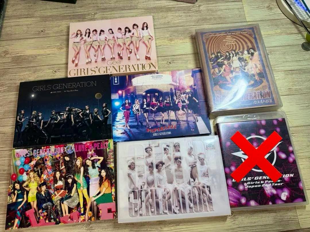 Several Girls' Generation CD, DVD, and Blu-ray cases are arranged on a wooden surface, with one case marked by a red X.