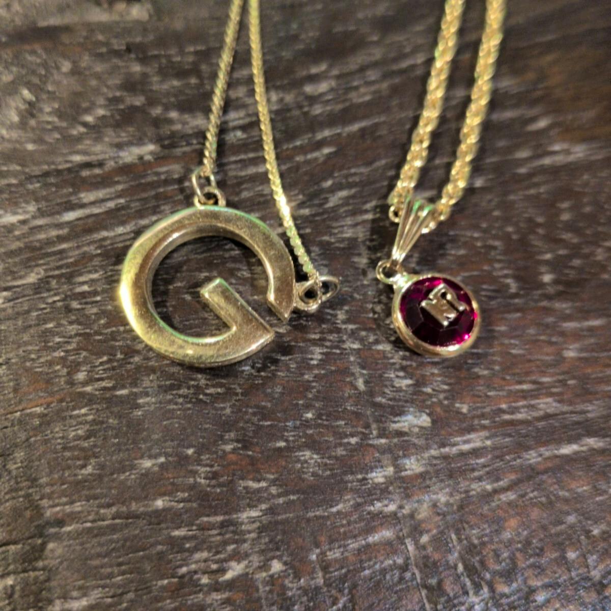 Two gold-tone necklaces with round pendants, one large 'G' pendant and one small red-stoned pendant, on a dark wooden surface.