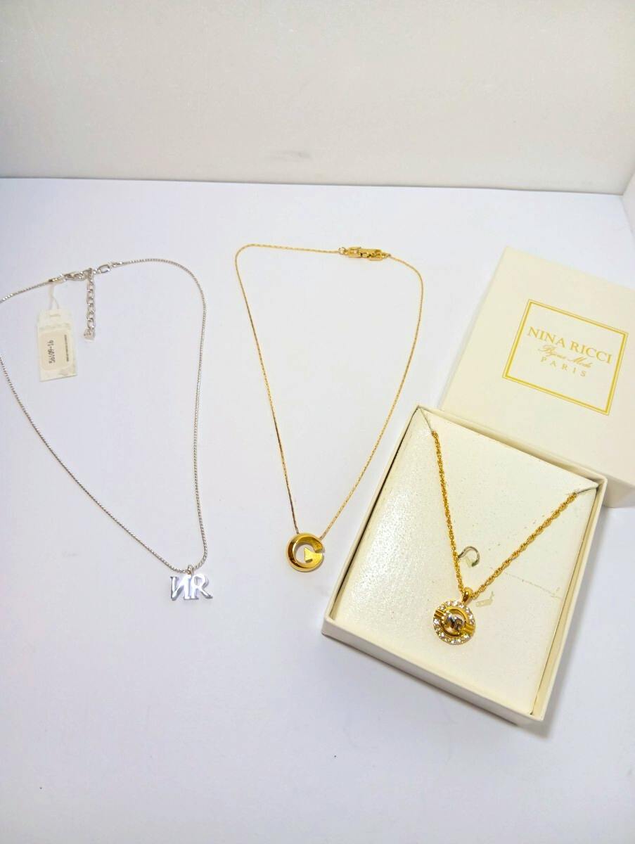 Three necklaces laid flat on a white surface, including a silver letter pendant, a gold disc pendant, and a gold necklace displayed in a Nina Ricci box.