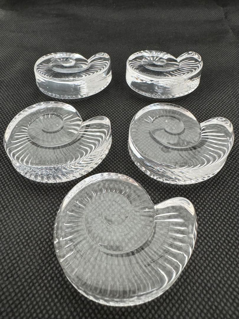 Five clear glass ammonite-shaped chopstick rests arranged on a dark textured surface.