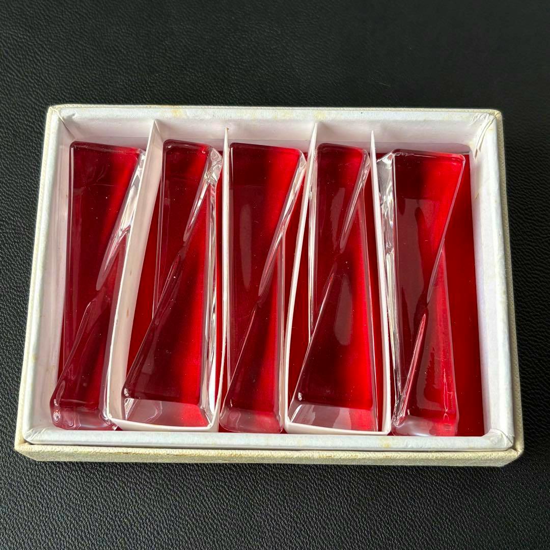 Five red glass chopstick rests displayed in a white partitioned box on a dark surface.