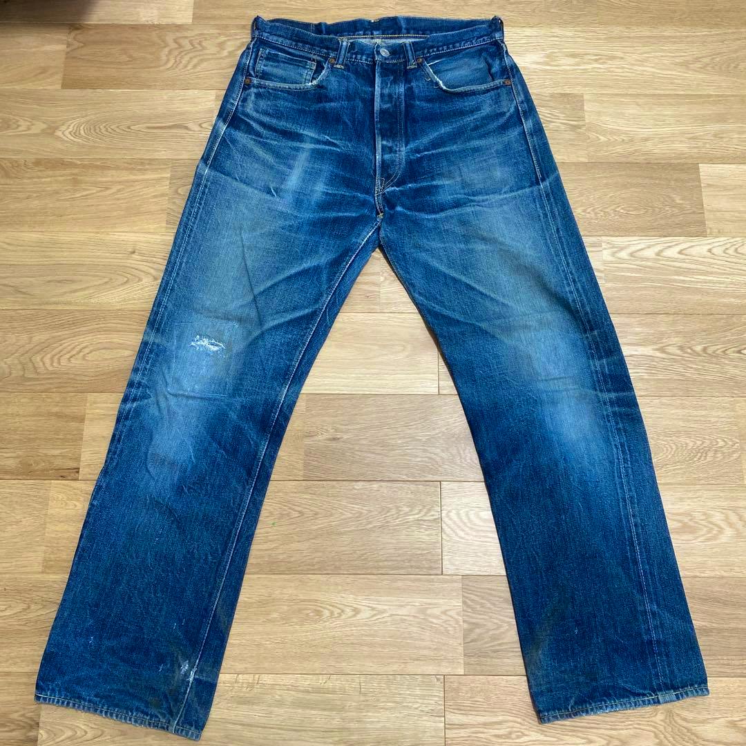 Flat-lay front view of men's blue denim jeans on a wooden floor showing fading and a small distressed area.