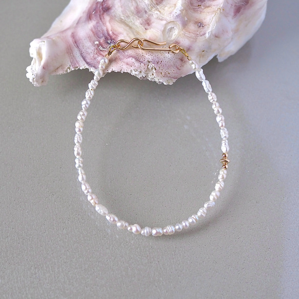 A baroque pearl bracelet with a gold infinity clasp laid flat on a gray surface with a shell prop behind it.