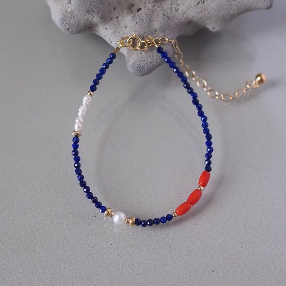 Close-up of a bracelet with dark blue lapis beads, red coral beads, a freshwater pearl, and a gold extension chain on a gray background.
