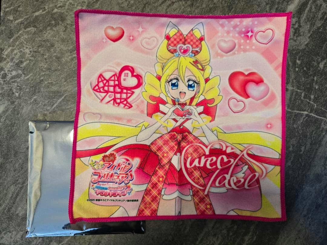 A square polyester handkerchief/towel printed with a pink anime-style girl and heart motifs, laid on a gray surface with a silver pouch visible at the lower left.