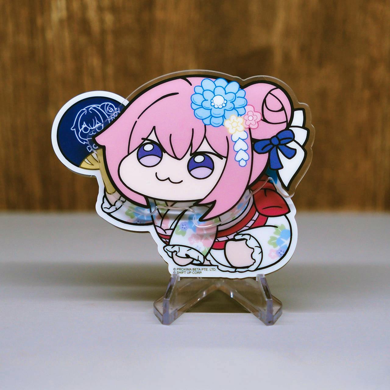 Photograph of the NIKKE "DORO" fridge magnet showing a pink-haired chibi character in a floral kimono displayed on a small clear stand.