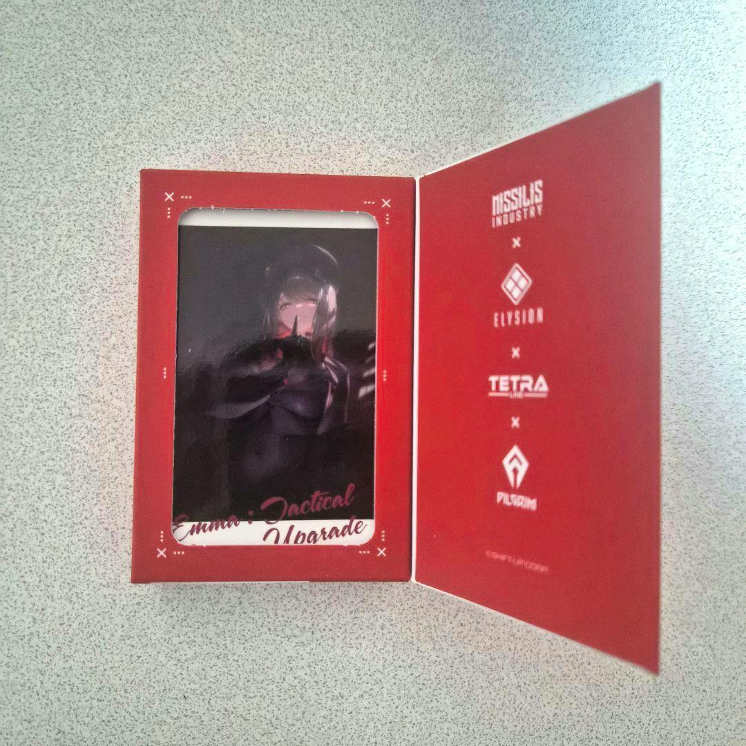Photocard for 勝利の女神NIKKE (隊員募集チョコレート vol.2 エマ) shown inside an open red fold-out sleeve on a light speckled surface.